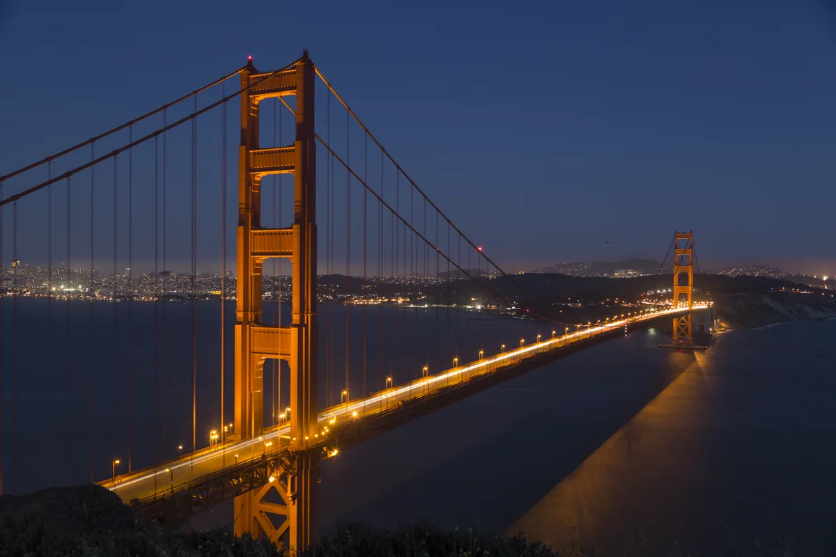The lit roadway and orange towers of the Golden Gate Bridge at dusk, with city lights in the distance