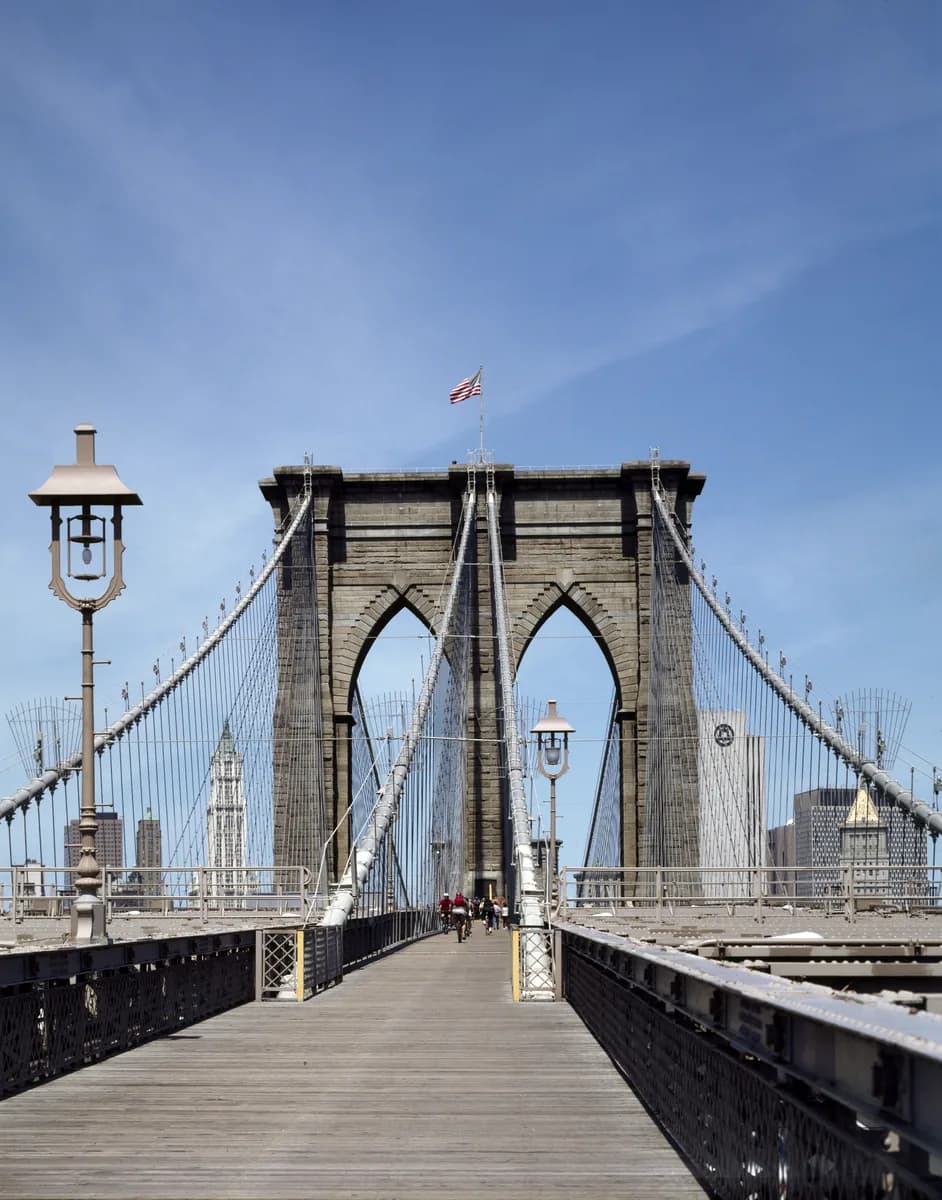 The stone tower and wire cables of the Brooklyn Bridge, seen from the wooden walkway under a blue sky