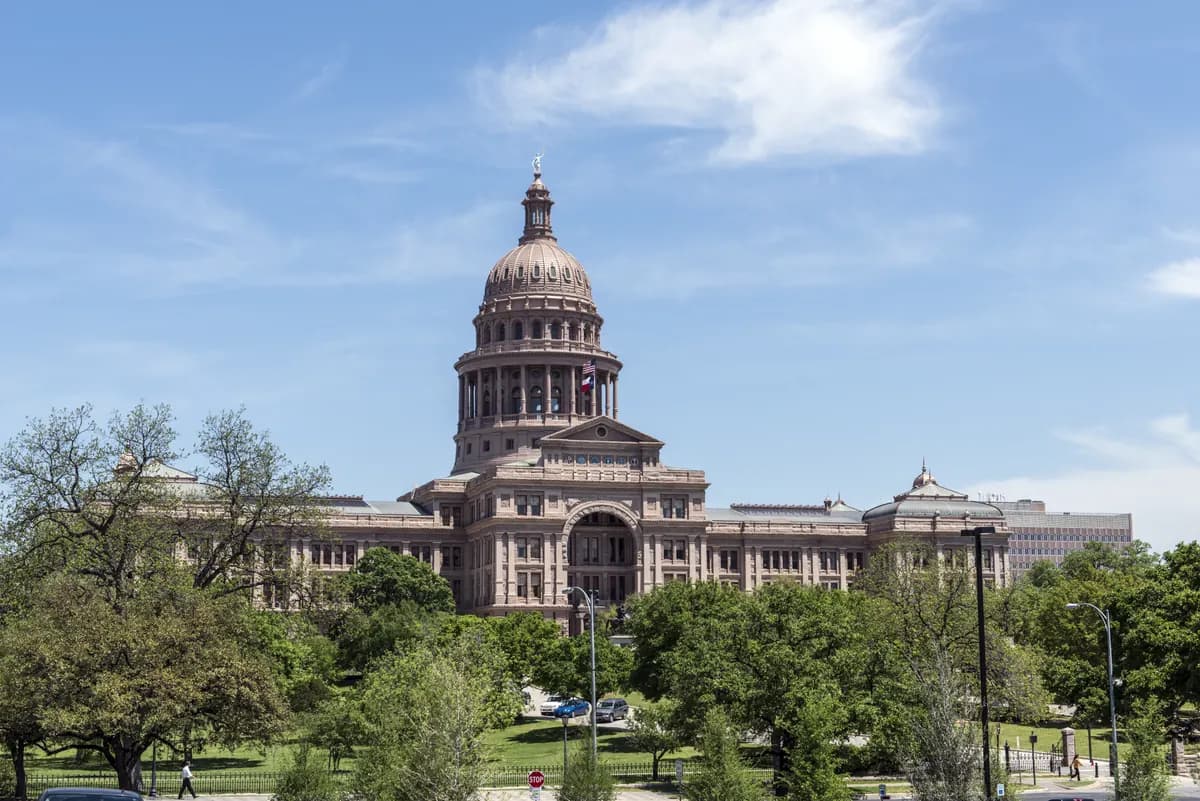 The Texas State Capitol in Austin, a pink granite building with a tall central dome, seen across green trees under a blue sky